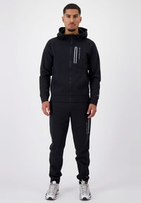 Zwarte zip-up hoodie met een voorzak en reflecterende accenten, gecombineerd met bijpassende zwarte joggers. Model draagt meerkleurige sportschoenen.