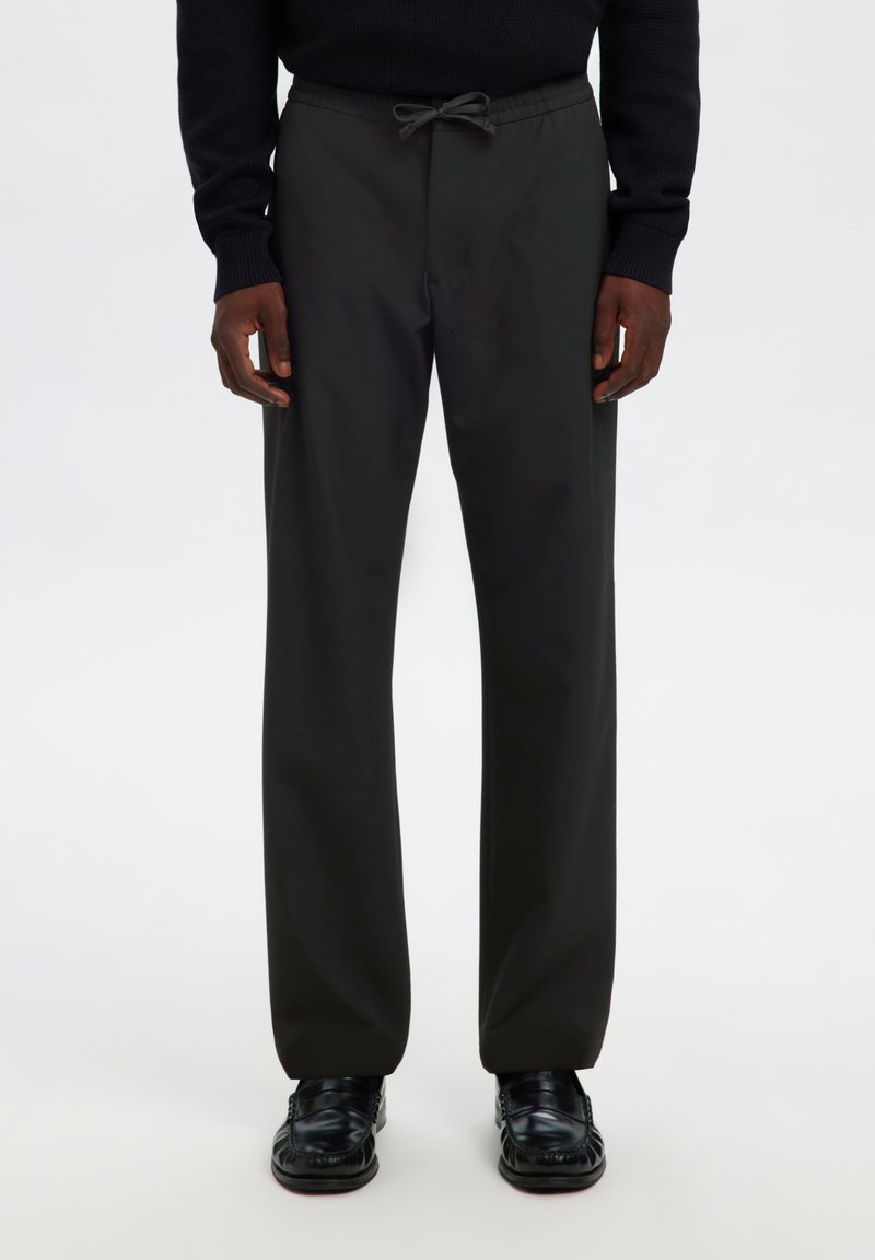 Hombre con pantalones negros holgados con cordón, combinados con zapatos de cuero negros y una camiseta negra de manga larga, de pie sobre un fondo blanco.