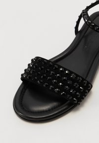 Svart sandal med flat såle, semsket rem dekorert med svarte krystallstifter, tynn ankelrem og glatt lærfôr.
