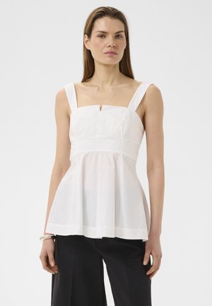 Vrouw staat, draagt een mouwloze witte peplum top met een vierkante halslijn en brede bandjes, gecombineerd met een zwarte broek tegen een effen achtergrond.