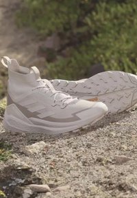 Jasnofioletowe buty trekkingowe z wysokim wsparciem kostki i chropowatymi podeszwami, umieszczone na skalistym terenie z rozmytym zielonym tle w tle.