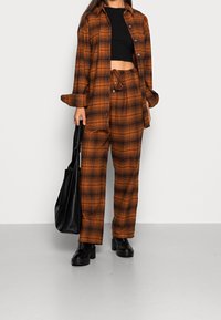 Camicia a quadri oversize marrone e nera abbinata a pantaloni coordinati, con vita regolabile e un top nero corto. Borsa tote nera e stivali.