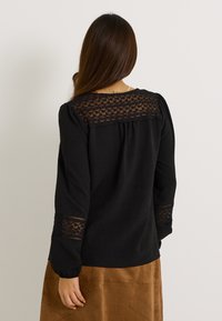 Blusa negra con detalles de encaje transparente en los hombros y las mangas, fabricada con una tela ligeramente texturizada. Manga larga y diseño de corte relajado.