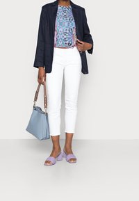 Blazer navy sobre una blusa estampada, jeans blancos, sandalias de tacón trenzadas moradas y un bolso azul claro con asa de cadena.
