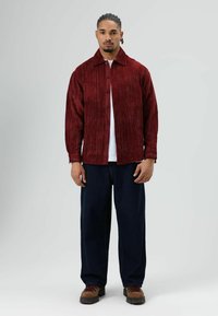 Rode corduroy blouse met knopen en kraag, gedragen over een wit T-shirt, gecombineerd met marineblauwe wijde broekspijpen en bruine veterlaarzen.