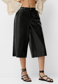 Svarta faux läder culotter med en mjuk yta, bred ben-design och dragkedja, kombinerat med svarta sandaletter med remmar och silverdetaljer.