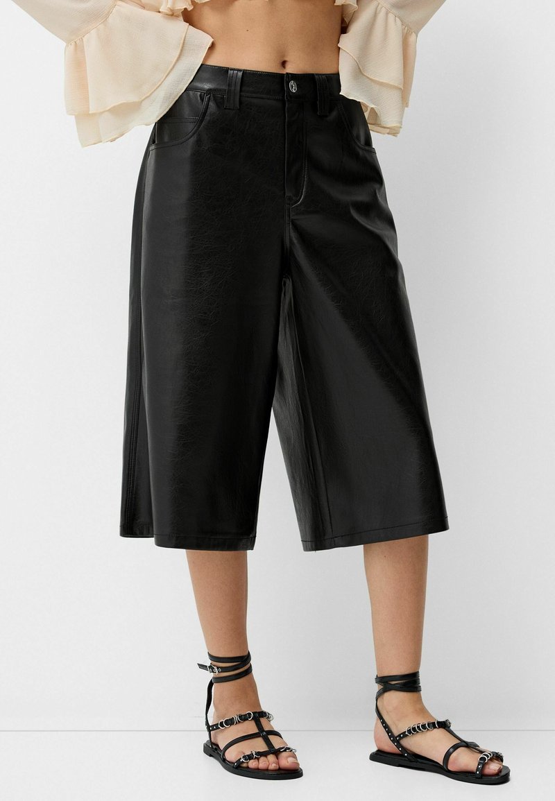 Svarta faux läder culotter med en mjuk yta, bred ben-design och dragkedja, kombinerat med svarta sandaletter med remmar och silverdetaljer.