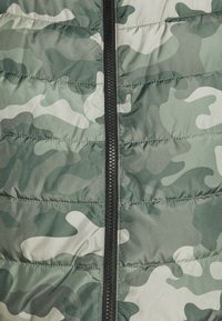 Tissu matelassé à motif camouflage vert et gris avec une fermeture éclair noire centrée qui court verticalement.