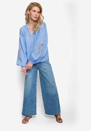 Lichtblauwe katoenen blouse met volumineuze mouwen en bloemrijke borduursels op de manchetten, gecombineerd met brede blauwe denim jeans en bruine sandalen.