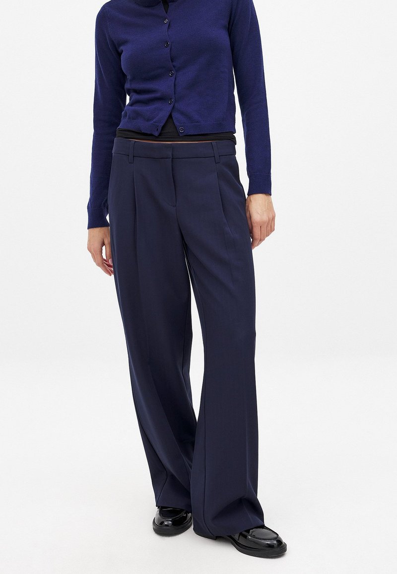 Navy weitgeschnittene Hose mit Falten, gefertigt aus glattem Stoff. Kombiniert mit einem navyfarbenen Knopf-Cardigan und schwarzen Schuhen.