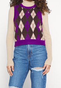 Lila och beige argylemönstrad stickad väst med hög krage, kombinerad med slitna blå jeans med hög midja. Texturerad, figurformad design.