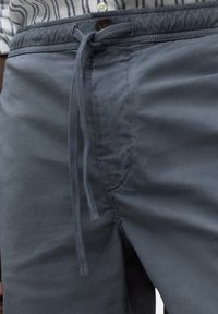 Pantalones cortos grises, ligeros, con cinturilla elástica, cierre de cordón y un bolsillo frontal. Textura suave con un brillo sutil.