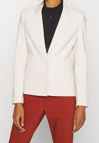 Blazer ajusté en crème clair avec revers, fermeture à un bouton et coupe structurée, associé à une chemise noire à col haut et un pantalon rouge.