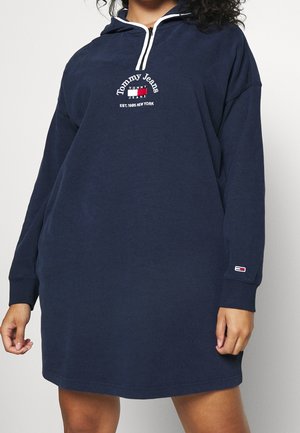 Kobieta w granatowej, długorękawowej sukience z kapturem, białym zamkiem błyskawicznym i logo Tommy Jeans na piersi i rękawie.
