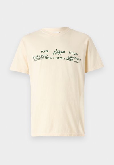 Kreminės spalvos medvilninis marškinėliai su trumpomis rankovėmis, ant kurių yra žalias užrašas "KidSuper", "Super Fluff n' Fold" ir skalbimo informacija.