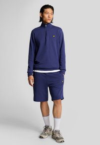 Marinblå kvartdrag sweatshirt med en liten gul logotyp, tillsammans med marinblå shorts. Modellen har vita strumpor och grå sneakers. Avslappnad aktiv stil.