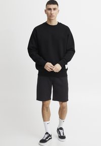 Sudadera de algodón negra con mangas largas, cuello redondo y puños acanalados. Combinada con pantalones cortos negros y zapatillas negras con detalles en blanco.
