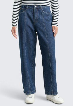 Un jean en denim bleu foncé taille haute avec une coupe évasée, des coutures contrastantes et des poches avant, associé à des baskets blanches.