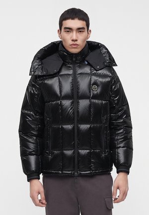 Junger Mann trägt eine glänzende schwarze Steppjacke mit hohem Kragen und Reißverschluss, steht vor einem schlichten hellen Hintergrund.