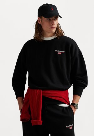 Man with long hair wears black Polo Sport sweatshirt and pants, black cap, red sweater tied around waist, and wristwatch, looking sideways.