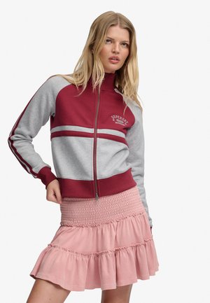 Blonde vrouw die een bordeauxrode en grijze jas met rits en strepen draagt, gecombineerd met een gestructureerde roze gerimpelde rok, poseert met één hand op de heup.