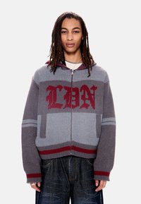 Άτομο με μακριές κοτσίδες dreadlocks που φοράει γκρι πλεκτό hoodie με φερμουάρ, με κόκκινες επιγραφές και ρίγες, συνδυασμένο με φαρδιά σκούρα τζιν παντελόνια.