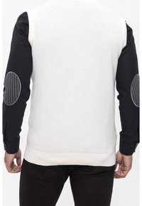 Chaleco de suéter blanco texturizado con un patrón de panal, combinado con una camisa negra de manga larga que tiene coderas de pata de gallo.