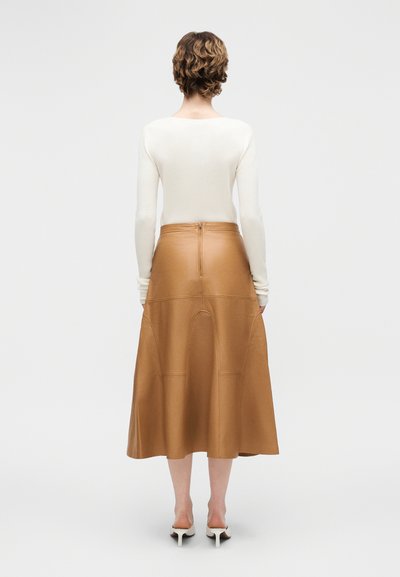 Άτομο με κοντά, σγουρά μαλλιά φοράει λευκό μακρυμάνικο τοπ, δερμάτινη μεσαίου μήκους φούστα σε καφέ ανοιχτό χρώμα με φερμουάρ στο πίσω μέρος και λευκά παπούτσια με τακούνι, ενώ στέκεται με την πλάτη προς τα έξω.