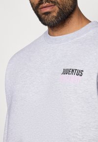 Felpa grigia chiaro con collo a giro. Presenta la scritta ricamata in nero e rosa "JUVENTUS" e "MILLE897" sul petto. Texture morbida.