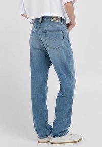 Person med ljusblå raka jeans och vita sneakers, stående med händerna i fickorna mot en enfärgad bakgrund.