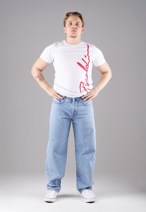 Weißes T-Shirt mit rographic auf der linken Seite, kombiniert mit hellblauen, lässig geschnittenen Jeans und weißen Sneakern. Minimalistisches Design, lässiger Look.