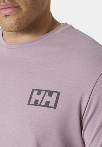 Camiseta de color lavanda claro hecha de tela suave, con un logo negro "HH" en el lado izquierdo del pecho. Cuello redondo y textura suave.