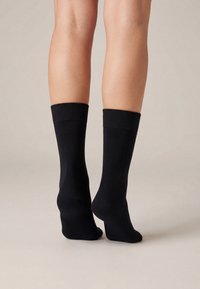 Noge u običnim crnim čarapama stoje na neutralnoj pozadini, pogled iz stražnje strane s jednom petom blago podignutom.