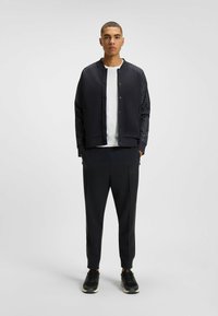 Black bomber jacket with patterned sleeves, white T-shirt underneath, black tapered pants, and black athletic shoes. Minimal design, casual fit.