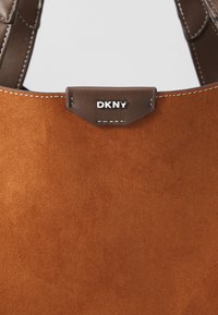 Bruine suede handtas met donkerbruin leren afwerking en witte stiksels, met een klein zilveren DKNY-logo plaatje.