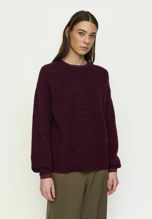 Femme aux longs cheveux bruns portant un pull ample bordeaux foncé et un pantalon vert olive, debout devant un fond clair uni.