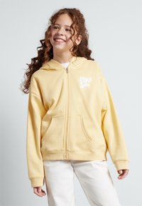 Sudadera amarilla con cremallera hecha de tela suave, que presenta un bolsillo frontal, puños acanalados y un logo blanco de "SURF" en la parte superior izquierda.