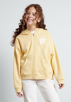 Haut zippé jaune en tissu doux, doté d'une poche frontale, de poignets côtelés et d'un logo "SURF" blanc sur le coin supérieur gauche.