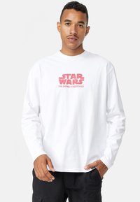 Valkoinen pitkähihaisesti T-paita, joka on valmistettu puuvillasta ja jonka edessä on punainen "Star Wars: Imperiumin vastaisku" -grafiikka.