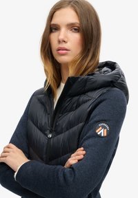 Chaleco acolchado negro con capucha sobre un suéter texturizado azul oscuro. Presenta una cremallera, un parche de logo en la manga y un cuello alto.