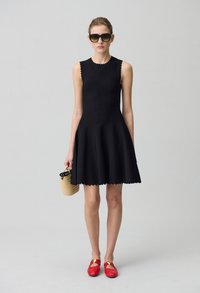 Robe noire sans manches avec un ourlet en forme de vagues, tissu texturé et découpes subtiles. Associée à des chaussures rouges et un petit sac en osier.