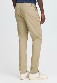 Beige cotton trousers with a relaxed fit, featuring back pockets, and worn with white athletic shoes. Smooth texture with minimal detailing.