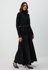 Un pull col roulé noir associé à une jupe maxi plissée noire, une ceinture dorée à imprimé léopard et des chaussures à talons noires. Texture lisse.