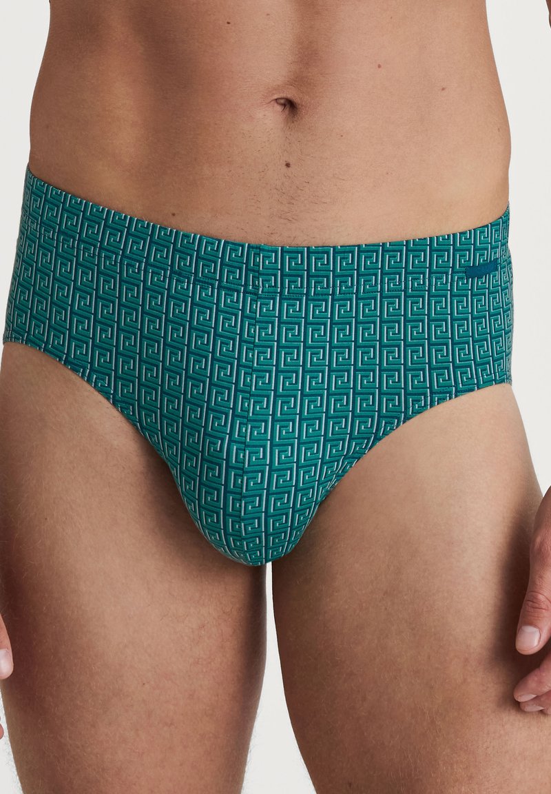 Türkisfarbene Schwimmslips mit einem geometrischen weißen Muster, ausgestattet mit einer glatten Textur und einem elastischen Bund für Komfort und Passform.