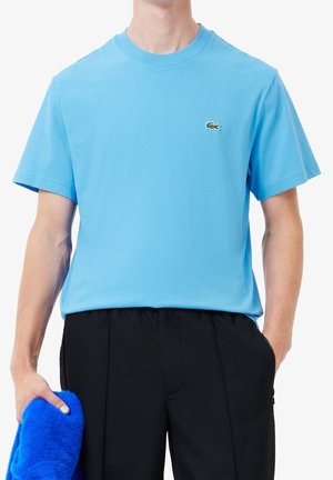 Mand iført en lyseblå Lacoste T-shirt og sorte bukser, der holder et lyst blåt, blødt tøjde i venstre hånd.