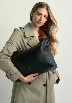 Kobieta o długich blond włosach w beżowym płaszczu trencz trzyma dużą czarną skórzaną torbę na jasnym tle.