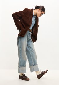 Jachetă din suede maro peste o cămașă din denim, asortată cu blugi strâmți albaștri. Pantofi maro și șosete de culoare crem completează ținuta.