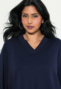Top de cuello en V en color navy, hecho de una tela suave; cuenta con mangas largas y una textura lisa. Pendientes geométricos en forma de aro destacan el look.