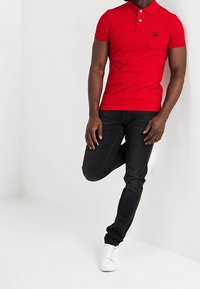 Rotes Polohemd mit Kragen und drei Knöpfen, mit einem kleinen schwarzen Logo auf der linken Brust. Kombiniert mit schwarzen Jeans und weißen Sneakers.