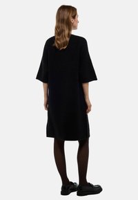 Robe tricotée noire avec une coupe ample, des manches tombantes et une longueur au genou. Assortie à des chaussures noires et des collants pour un look complet.